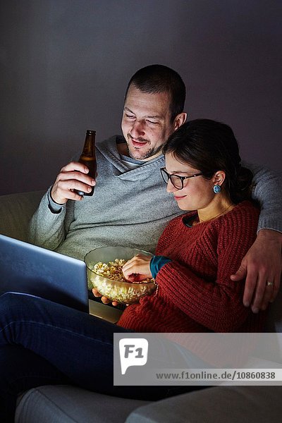 Mid adult couple sitting on sofa in evening eating popcorn and looking at laptop