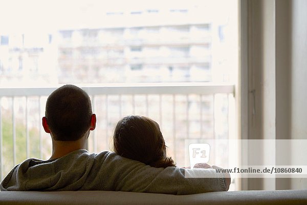 Rückansicht eines auf einem Sofa sitzenden Paares  das aus dem Fenster auf ein Wohnhaus schaut