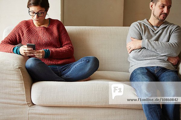 Schmollender Mann sitzt auf Sofa  während seine Freundin eine SMS liest