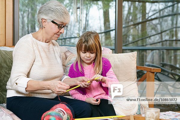 Ältere Frau unterrichtet Enkelin beim Stricken auf Wohnzimmersofa
