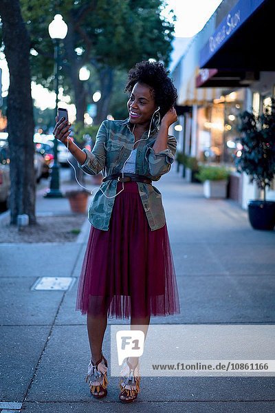 Frau steht auf der Straße  hält Smartphone  trägt Kopfhörer  lächelt