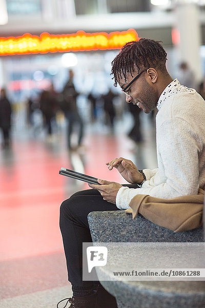 Junger Mann sitzt im Bahnhof und benutzt digitalen Tablet-Touchscreen