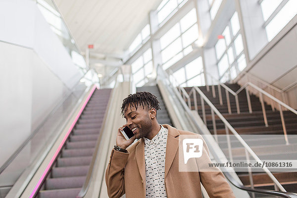 Junger Geschäftsmann spricht auf Smartphone und fährt die Rolltreppe des Bahnhofs hinunter