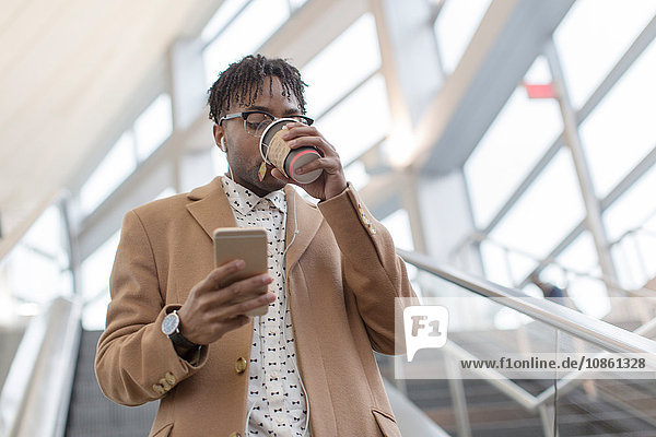 Junger Geschäftsmann fährt die Rolltreppe des Bahnhofs hinunter und trinkt Kaffee zum Mitnehmen