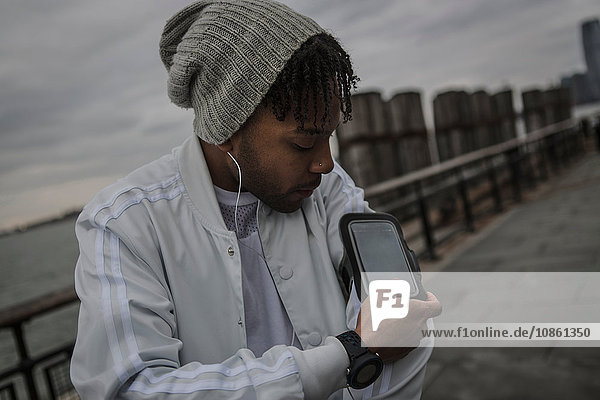 Junger Mann trainiert im Morgengrauen am Flussufer am Touchscreen eines Smartphones