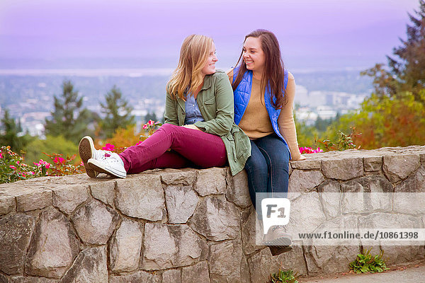 Zwei junge Frauen  die lächelnd von Angesicht zu Angesicht an der Wand sitzen