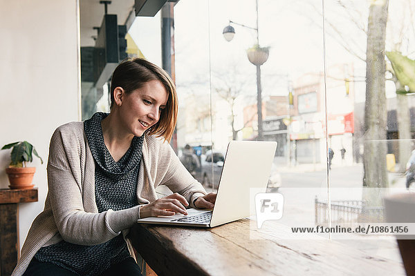 Female customer sitting in coffee shop window seat typing on laptop