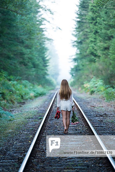 Teenagerin  geht an einem Zuggleis entlang  hält rote Schuhe und Blumen in der Hand  Rückansicht