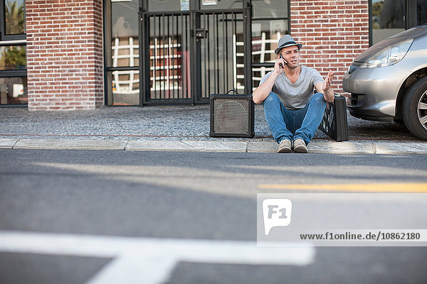 Straßenmusikant  der auf dem Bordstein sitzt und ein Mobiltelefon benutzt