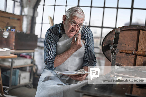 Senior-Handwerker spricht auf Smartphone in einer Antiquitäten-Restaurierungswerkstatt