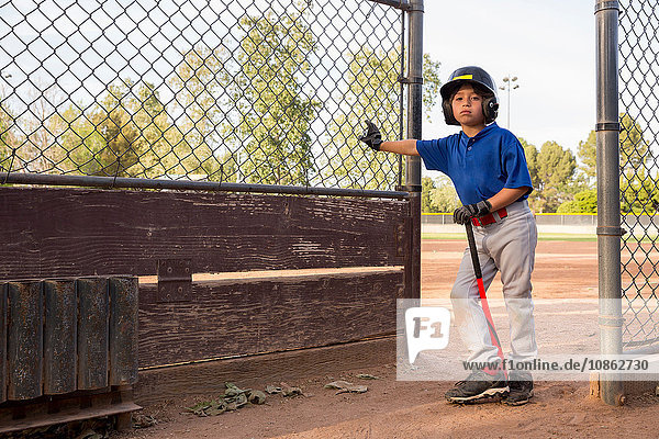 Porträt eines Jungen mit Baseballschläger  der sich beim Baseballtraining gegen den Zaun lehnt