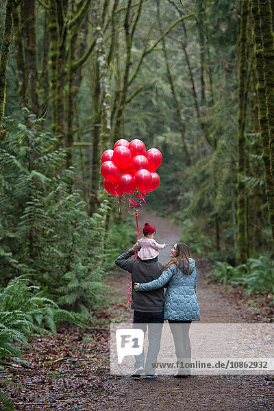 Rückansicht von Eltern  die mit der Kleinkind-Tochter auf den Schultern und einem Haufen roter Luftballons im Wald spazieren gehen