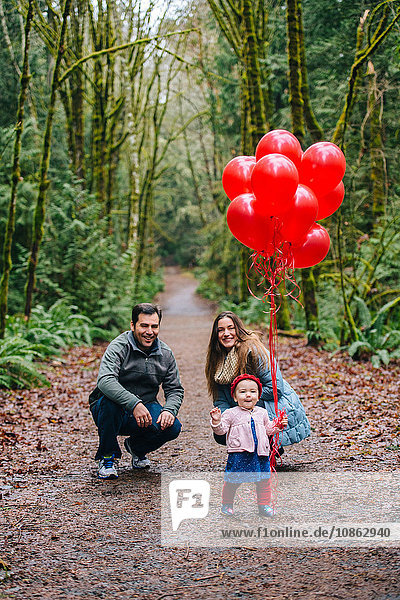 Porträt von Eltern  die ihre Kleinkind-Tochter mit einem Bündel roter Luftballons im Wald hocken