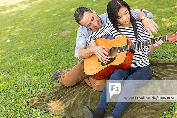 Mann sitzt auf Gras und lehrt Frau Akustikgitarre spielen