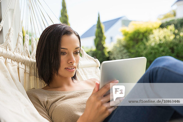 Junge Frau liegt auf Gartenhängematte und stöbert auf digitalem Tablet