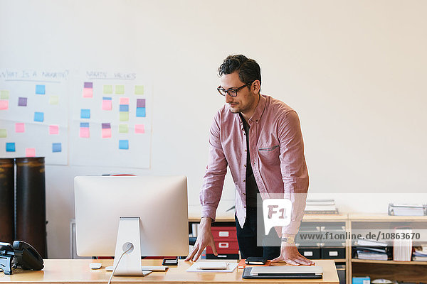 Mann im Büro lehnt am Schreibtisch und schaut auf den Computermonitor