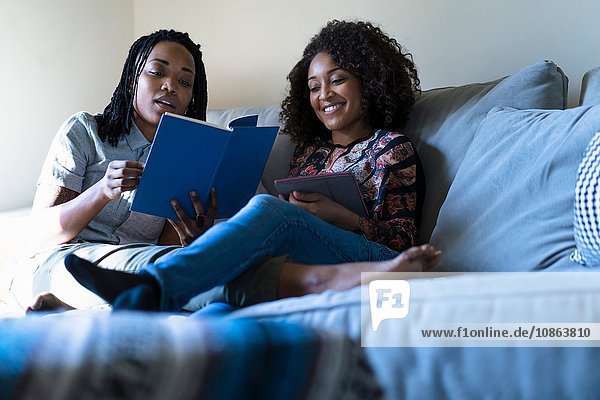 Lesbisches Paar entspannt sich auf dem Sofa  liest ein Buch und benutzt ein digitales Tablett