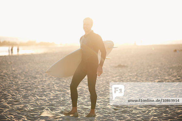 Male surfer carrying surfboard on sunlit Venice Beach  California  USA