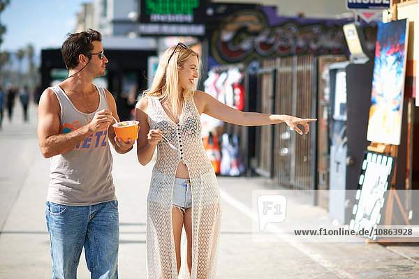 Couple eating frozen yoghurt and pointing at shops on sidewalk  Venice Beach  California  USA