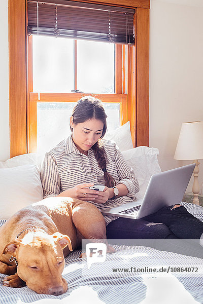 Frau sitzt mit Hund und Laptop mit Smartphone auf dem Bett