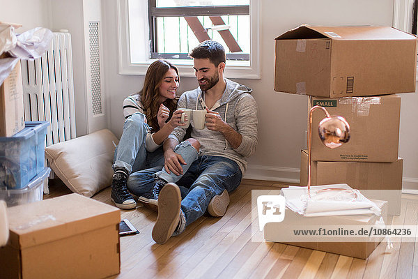 Moving house: Young couple sitting in room full of boxes  drinking hot drink