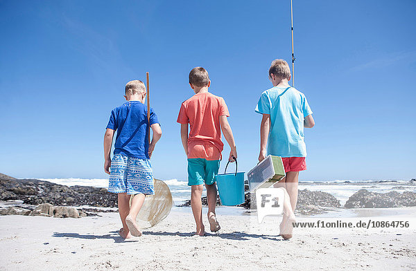 Rückansicht von drei Jungen,  die mit Fischernetz,  Eimer und Angelrute am Strand spazieren gehen,  Kapstadt,  Südafrika