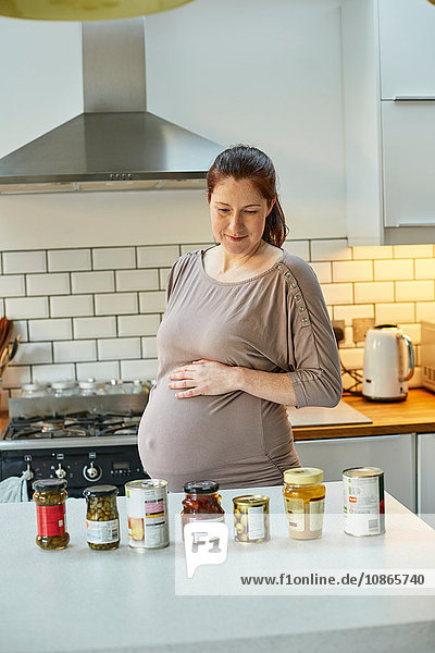 Schwangere Frau in der Küche schaut auf Gläser und Dosen mit Lebensmitteln auf der Küchentheke