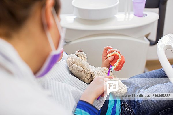 Schrägansicht eines Jungen auf einem Zahnarztstuhl  der einen Teddybären hält  der Zahnprothesen bürstet