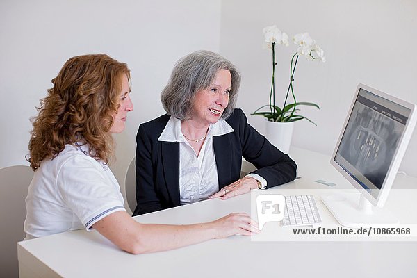 Ältere Frau und reife Frau am Schreibtisch diskutieren lächelnd über das Röntgenbild auf dem Computerbildschirm