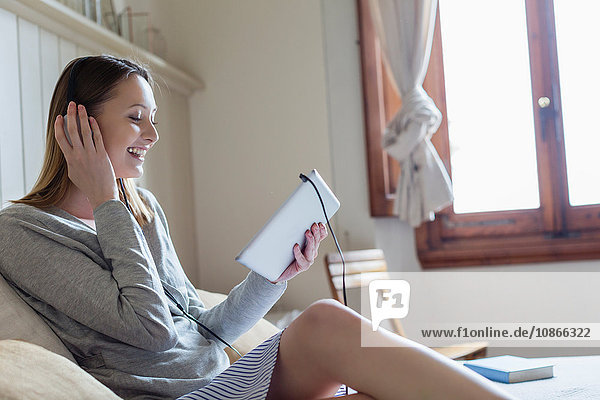Frau sitzt mit Kopfhörer auf dem Bett und schaut lächelnd auf das digitale Tablet