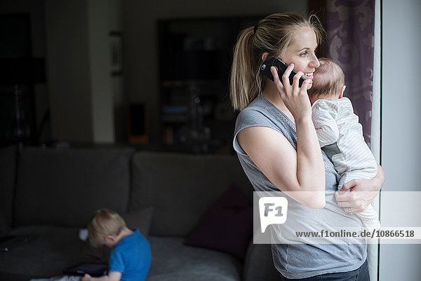 Mutter hält Baby Junge  spricht mit Smartphone  während Sohn digitales Tablet benutzt