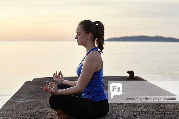 Young woman sitting on pier  in yoga position  at sunset