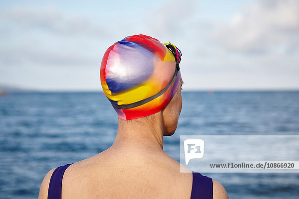 Mature woman wearing swimming costume and swimming hat  standing beside the sea  rear view