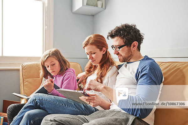 Mid adult parents and daughter on sofa concentrating on reading book and digital tablet