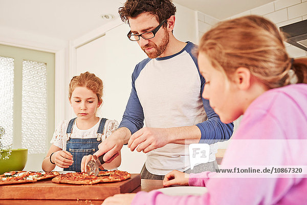 Mid adult man slicing pizza for daughters at kitchen bench