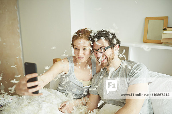Mittelgroßes  mit Kissenschlachtfedern bedecktes erwachsenes Paar nimmt Smartphone-Selfie im Bett