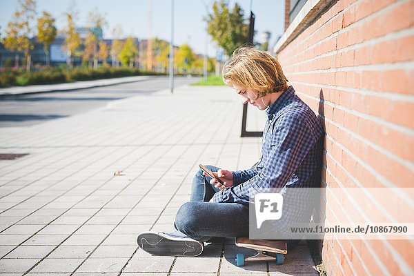 Junge männliche städtische Skateboarder sitzen auf dem Bürgersteig Auswahl Smartphone-Musik