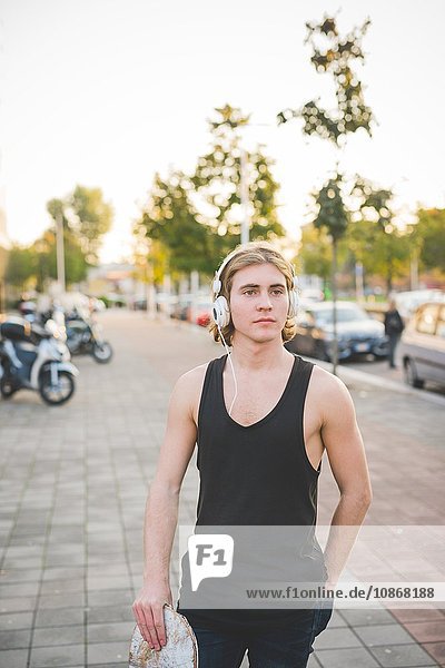 Portrait eines jungen männlichen Skateboardfahrers  der Kopfhörermusik auf dem Bürgersteig hört.