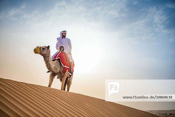 Man wearing traditional middle eastern clothes riding camel in desert  Dubai  United Arab Emirates