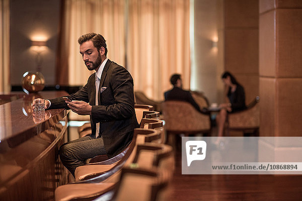 Junger Geschäftsmann sitzt an Hotelbar und liest Smartphone-Texte