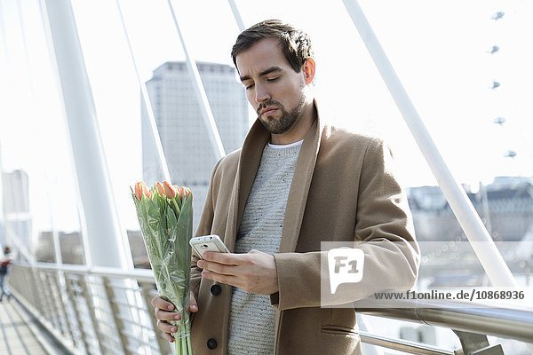 Mittelgroßer erwachsener Mann steht auf der Brücke  hält Blumen und schaut auf ein Smartphone