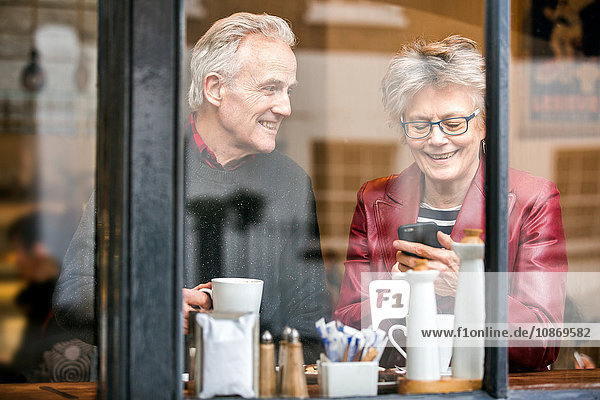 Älteres Ehepaar am Fensterplatz eines Cafés  trinkt Kaffee und schreibt SMS auf Smartphone