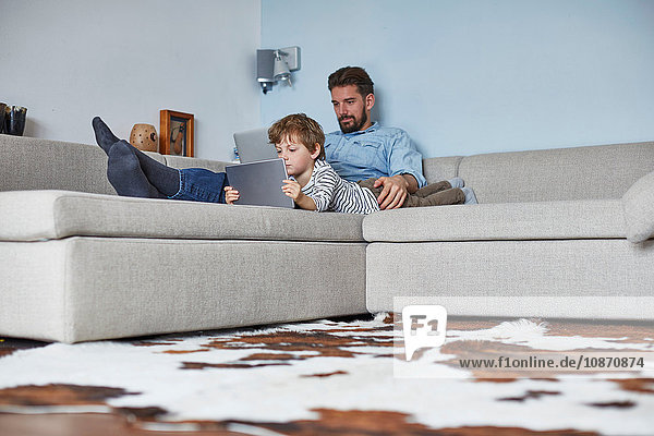 Boy lying on sofa with father using technology