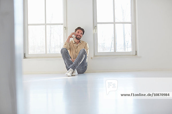 Junger Mann sitzt auf dem Boden und unterhält sich mit einem Smartphone in einem leeren neuen Haus