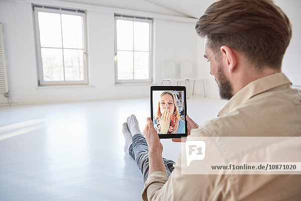 Girlfriend on digital tablet video call blowing a kiss to young man sitting on floor