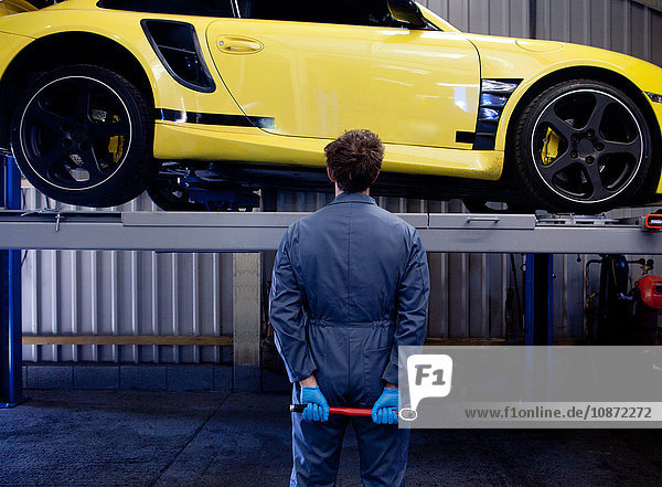 Male mechanic checking yellow sports car holding socket wrench behind his back rear view