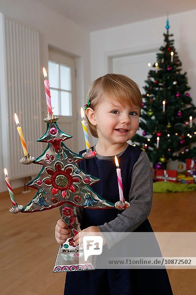 Mädchen vor dem Weihnachtsbaum hält Weihnachtsschmuck und schaut lächelnd in die Kamera