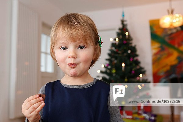 Mädchen vor dem Weihnachtsbaum isst Schokolade und schaut in die Kamera
