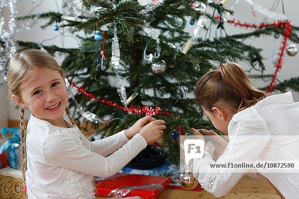 Mädchen schmücken Weihnachtsbaum mit Lametta und schauen lächelnd über die Schulter in die Kamera