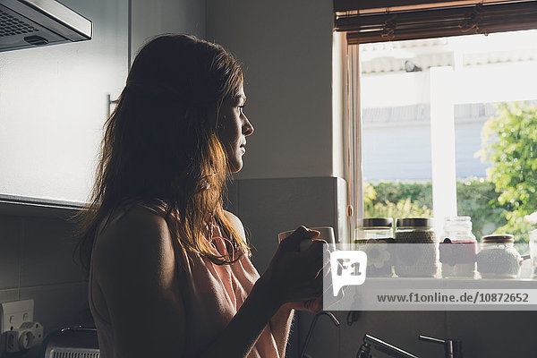 Junge Frau bei einer Kaffeepause  die durch ein Küchenfenster blickt